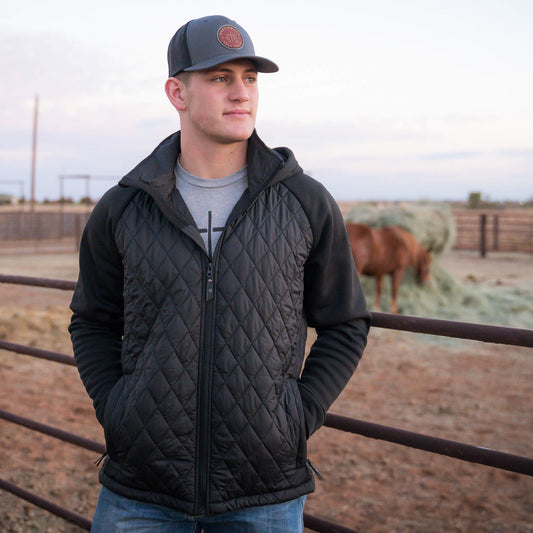 Man leaning against a metal fence on a ranch wearing a gray hat and black quilted jacket with a hood, jersey knit sleeves, and zip closure.