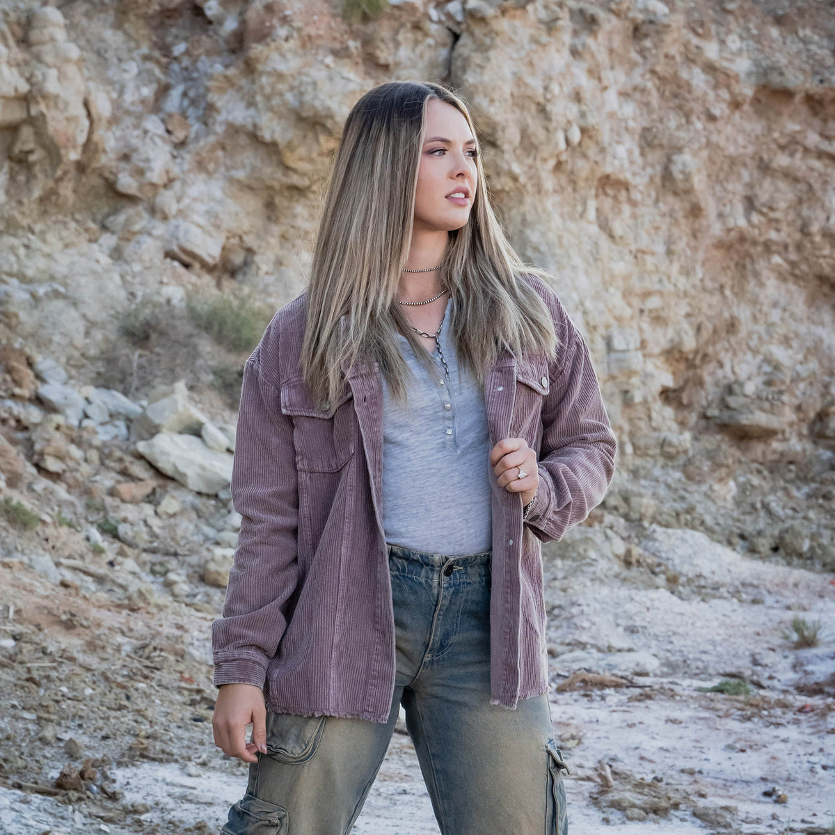 Woman standing in a rocky desert landscape wearing a purple corduroy jacket.