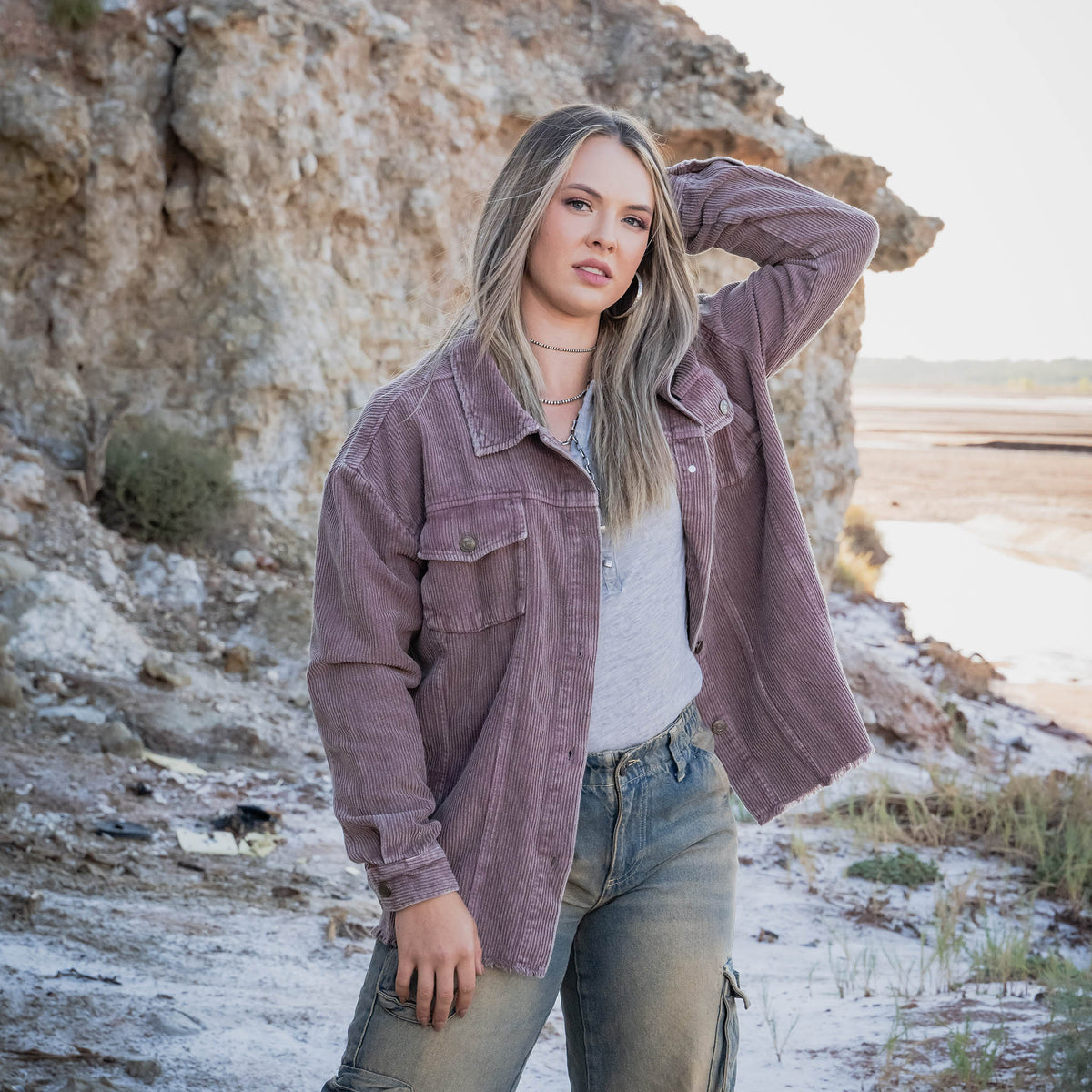 Woman in a purple corduroy jacket and jeans standing in a natural setting with rocks and a path.
