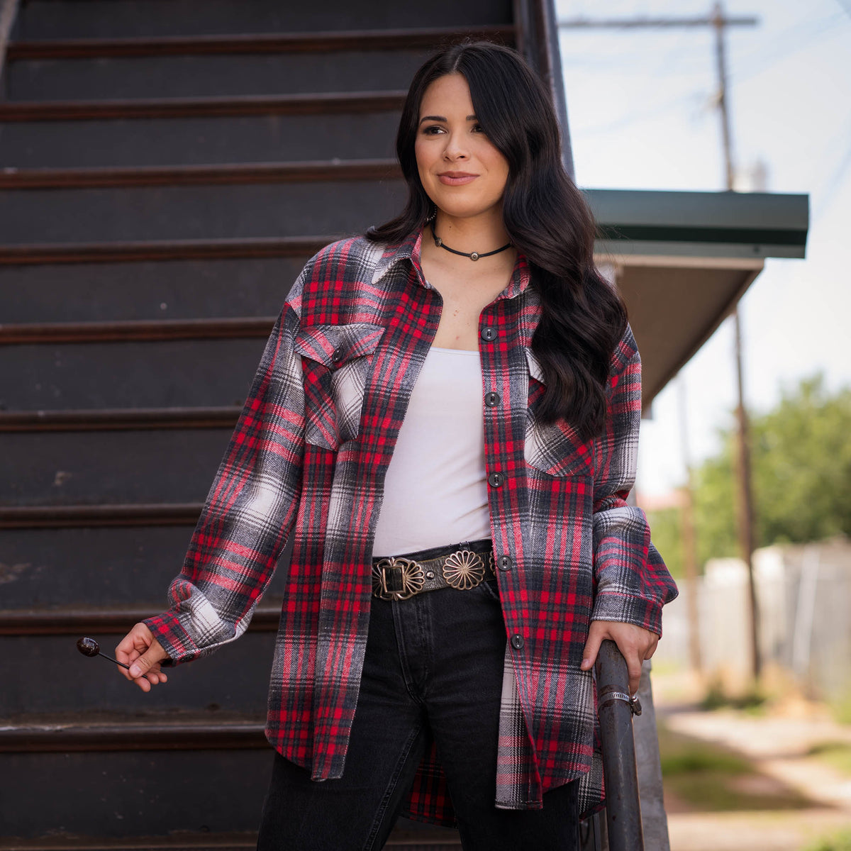 Woman standing on stairs wearing a white t-shirt, black jeans and a red, black, and white plaid women's shacket with button closures and chest pockets.