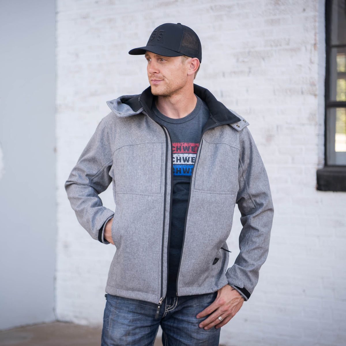 Man in front of brick wall wearing a black hat, jeans and heather gray softshell jacket with zip front closure, removable hood, and multiple pockets.