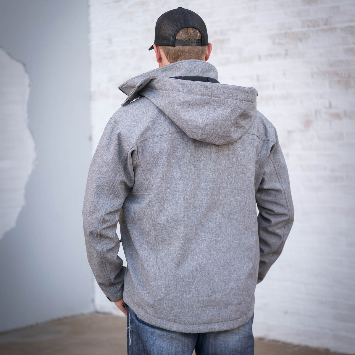 Man facing away in front of a brick wall wearing a heather gray softshell jacket with removable hood.