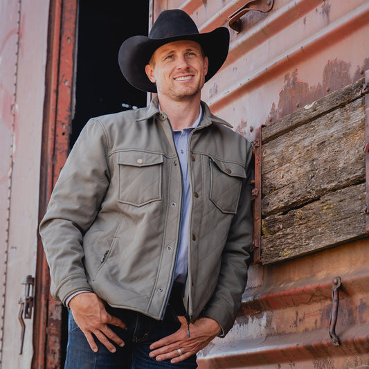 Clif Cooper outside sliding metal doors wearing our olive green jacket and black cowboy hat.