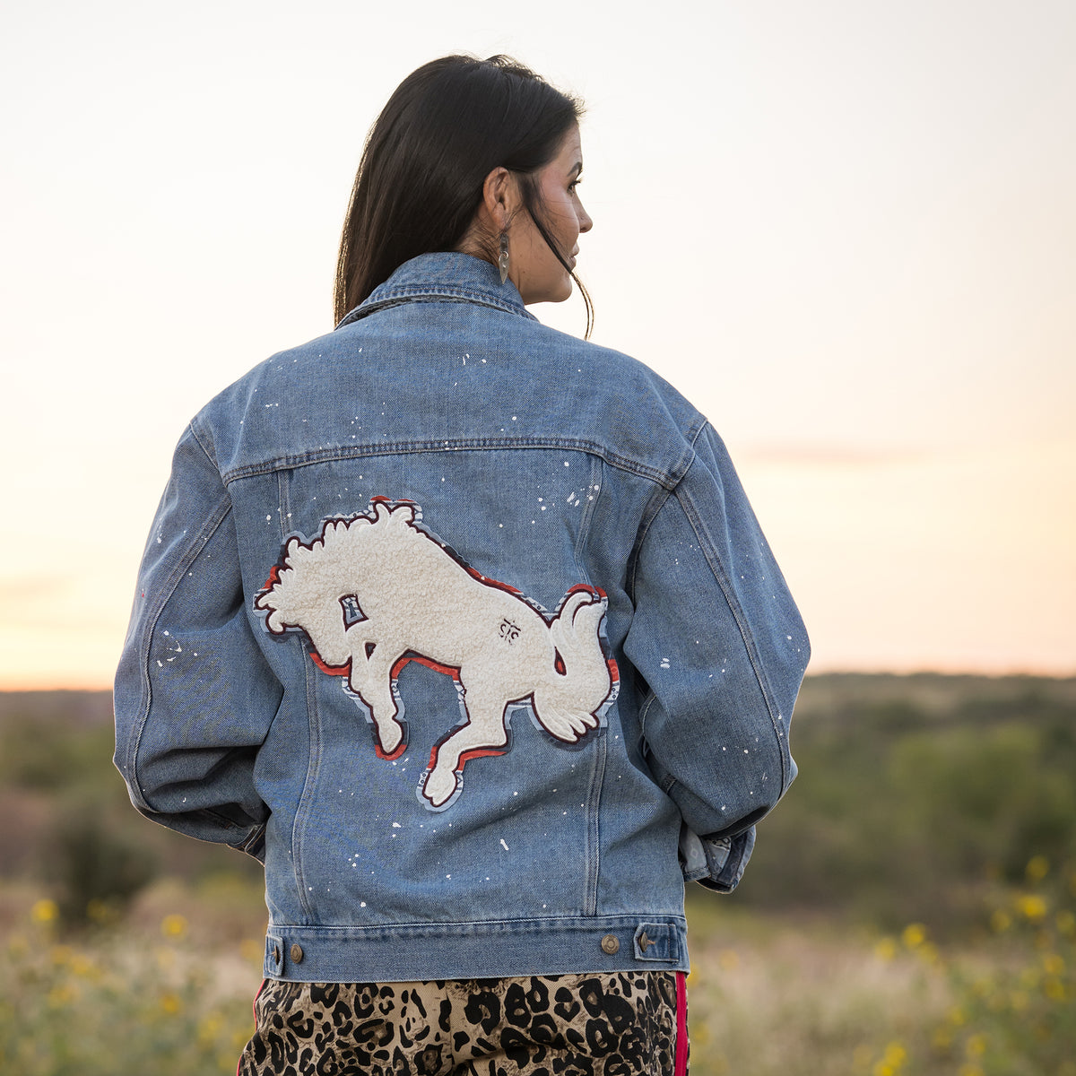 Woman facing away in a field wearing a spackled denim jacket featuring a chenille back patch.