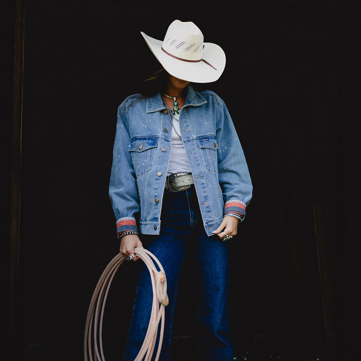 A person wearing a white cowboy hat, a light blue denim jacket stands against a dark background while holding a coiled rope.
