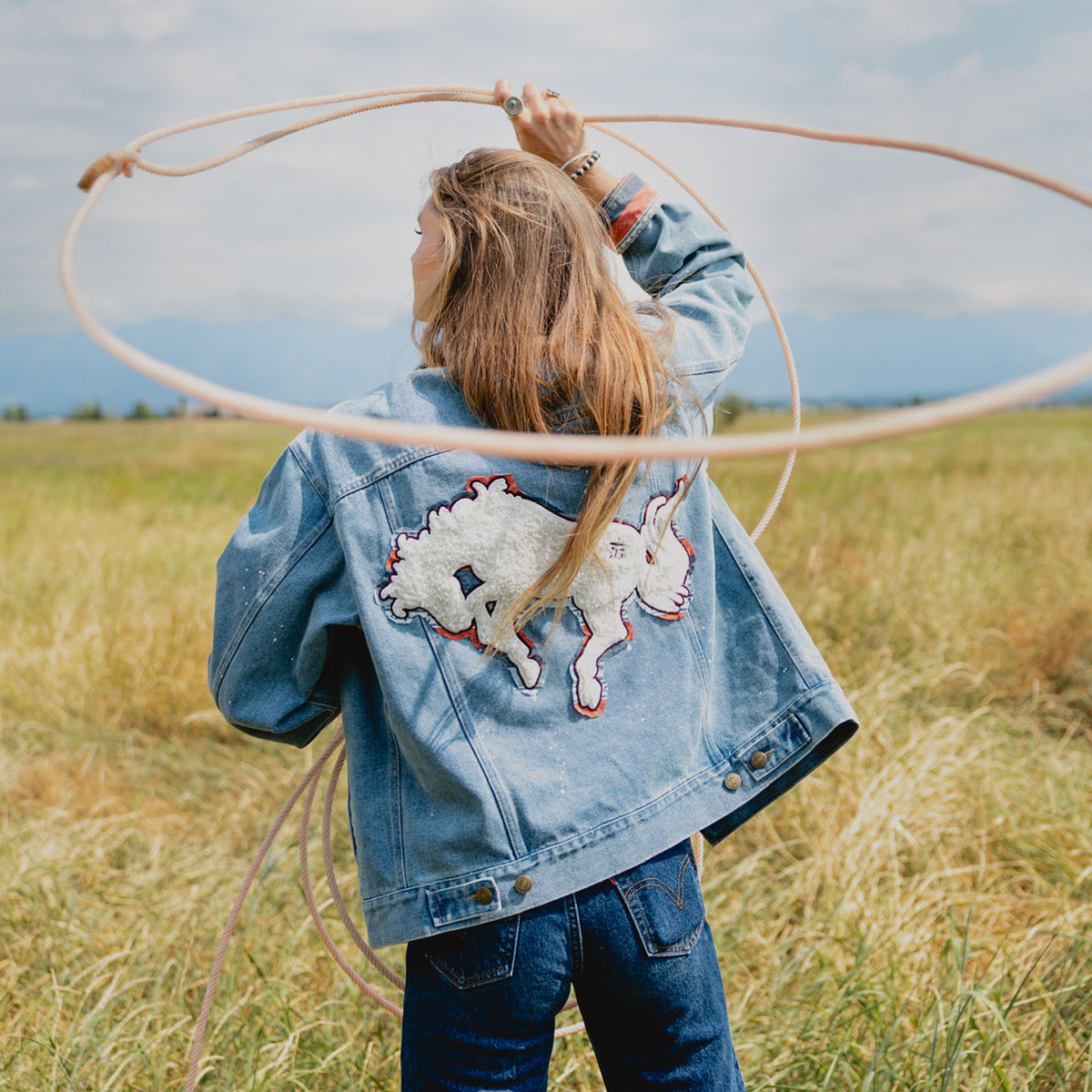 A woman stands in a grassy field with mountains in the background, viewed from behind. She is wearing a light blue denim jacket with a chenille patch on the back and holding a lasso overhead.