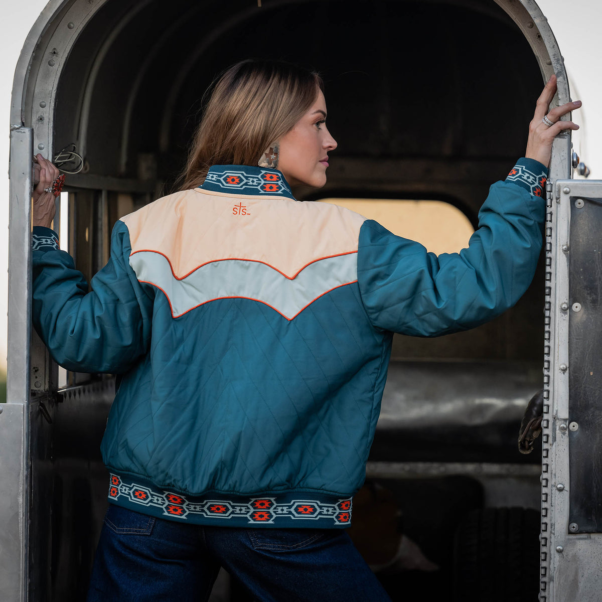 Woman wearing a turquoise and beige jacket with decorative patterns, standing inside a horse trailer.