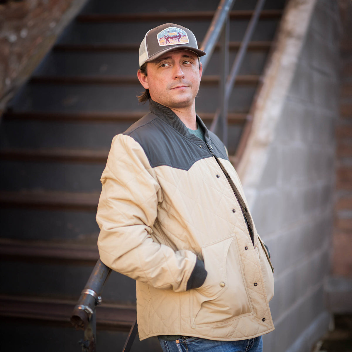 Mean leaning against a hand railing wearing a beige and black quilted jacket with double western yoke accents, ribbed collar and cuffs, and snap front closure.