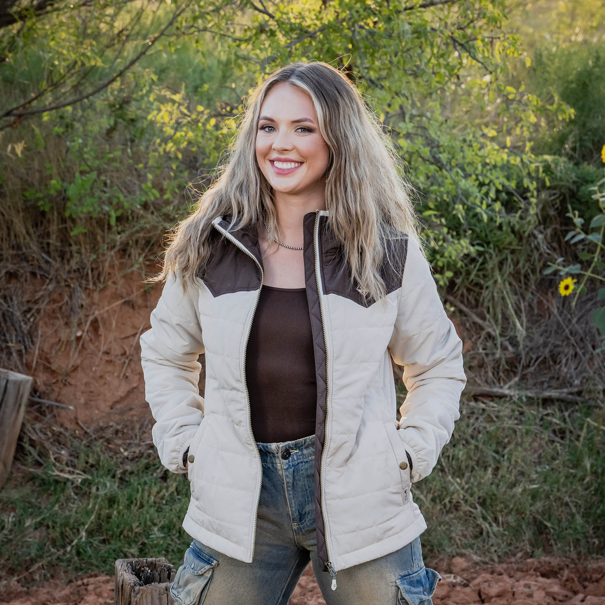Woman wearing a beige and brown puffer jacket outdoors with greenery in the background.