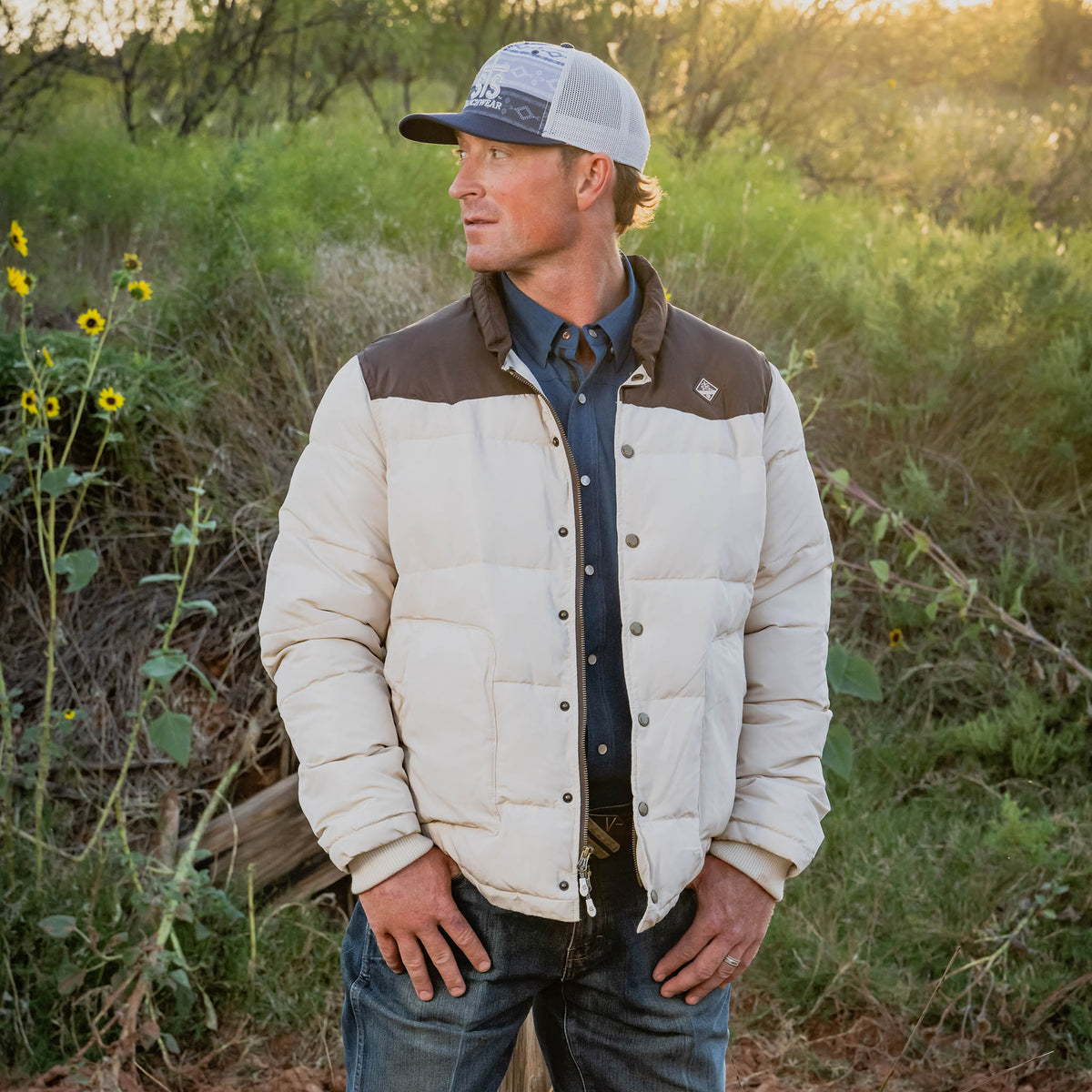 Man wearing a beige and brown jacket with a cap in a natural setting.
