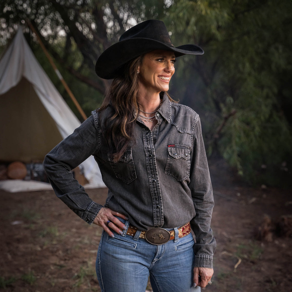 Jen Smith wearing a black cowboy hat, jeans and a women's pearl snap denim shirt with double western yoke and pearl snap chest pockets, in a dark wash in a forest setting.