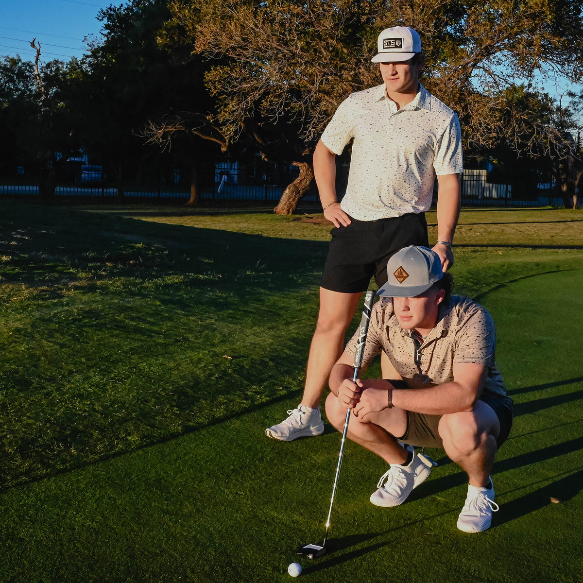Two men on a golf course standing behind a golf ball holding a putter.