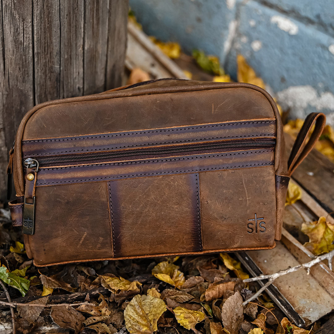 A brown leather Foreman Shave Kit bag with zip closure and the STS Ranchwear logo on the side sitting on leaves on the ground.