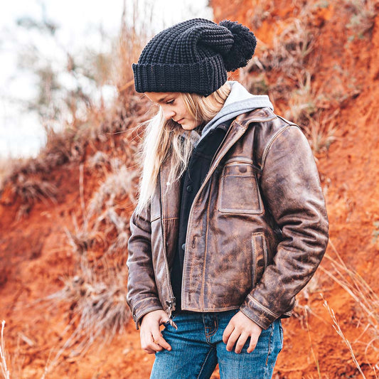 Young lady standing in a dirt field wearing a brown genuine leather jacket, black beanie and blue jeans.