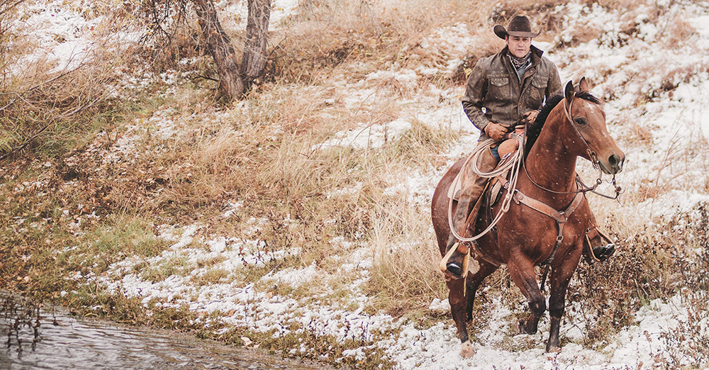 Stran T Smith wearing a cowboy hat and brown leather jacket riding a horse next to a creek in a snowy field.