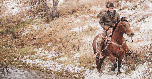 Stran T Smith wearing a cowboy hat and brown leather jacket riding a horse next to a creek in a snowy field.