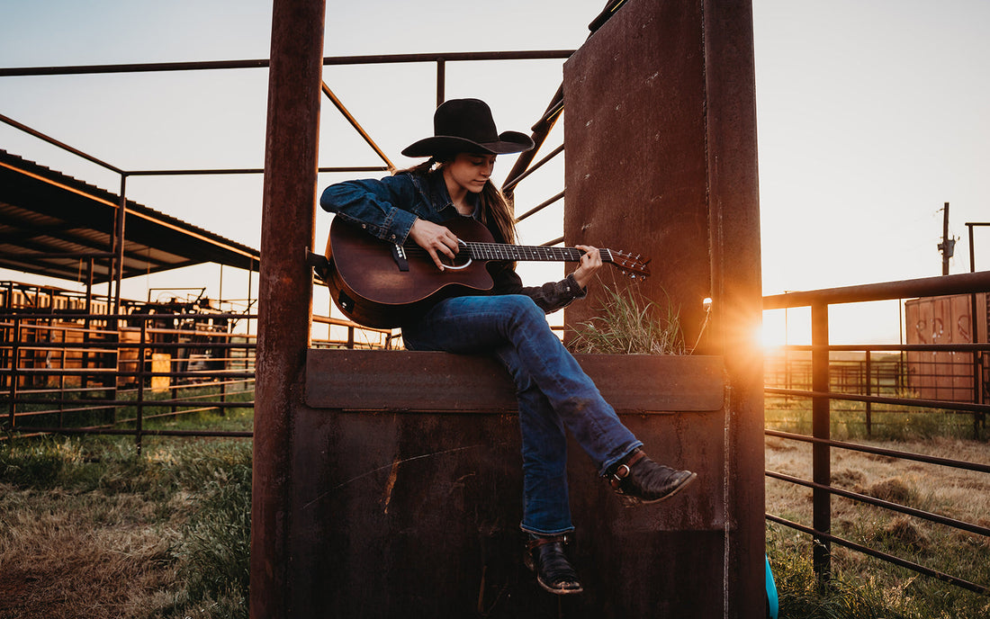 Selah Smith sitting on a wooden fence in front of a metal gate strumming a guitar in front of the sunset.