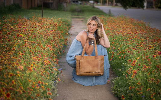 Woman wearing a blue denim dress kneeling on a sidewalk holding a tan leather tote bag surrounded by orange flowers.