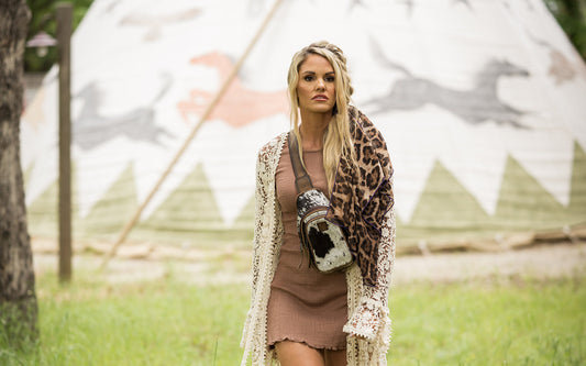 Woman wearing a tan dress with a hair on hide cowhide sling across their body standing in front of a teepee. 
