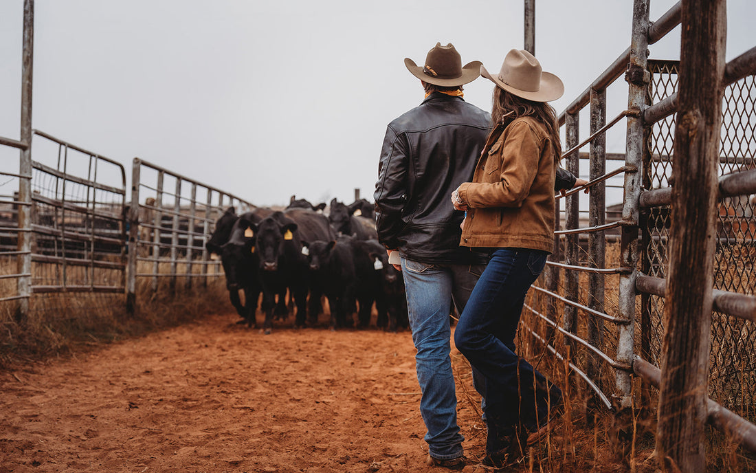 Stran and Jen Smith leaning against a metal gate standing in front of their Black Angus cattle on their ranch in Tell, Texas.