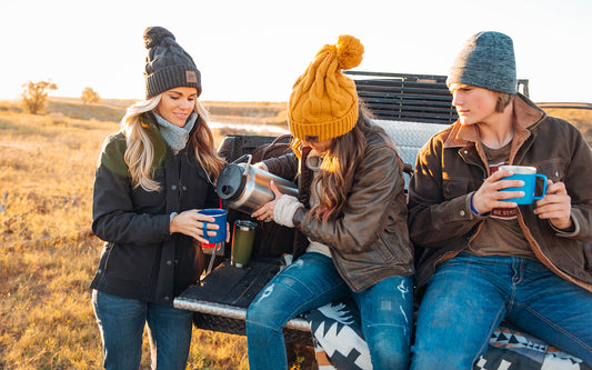 Three people sitting on a truck tailgate drinking hot chocolate in the middle of a grassy field.