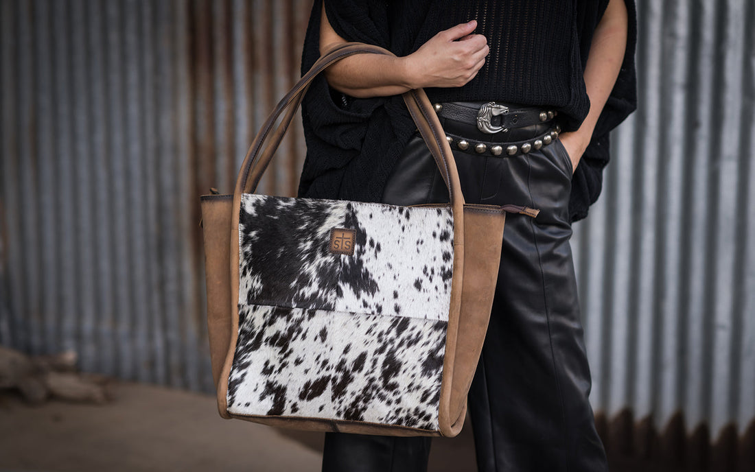 Person wearing all black holding a leather and hair on hide cowhide tote on their arm standing in front of a metal wall.