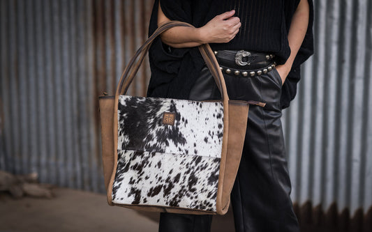 Person wearing all black holding a leather and hair on hide cowhide tote on their arm standing in front of a metal wall.