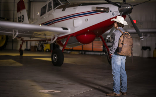 Man wearing a cowboy hat, gray shirt and blue jeans holding a brown leather backpack standing in front of a small airplane.