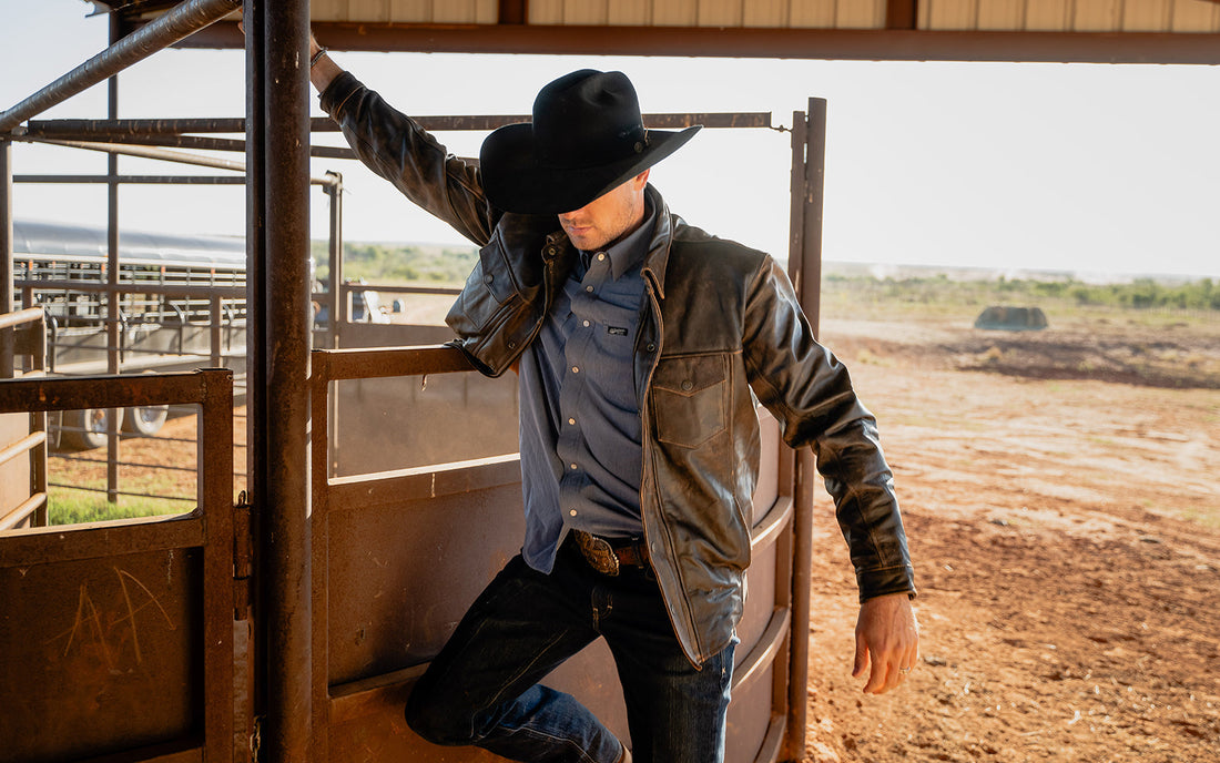 Man wearing a black cowboy hat and brown leather jacket leaning against a pole on a ranch in Tell, Texas.