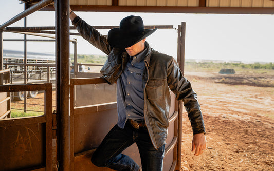 Man wearing a black cowboy hat and brown leather jacket leaning against a pole on a ranch in Tell, Texas.