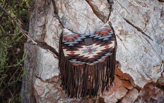 Brown, cream and gray hand woven shoulder bag with leather fringe hanging from a rock.