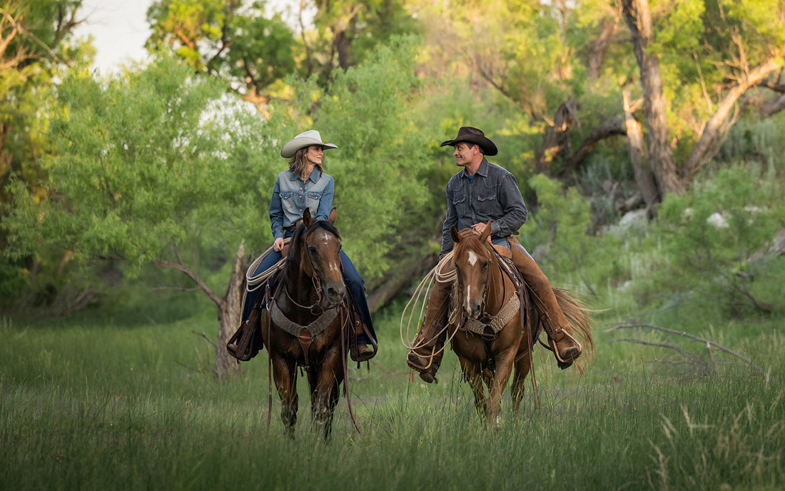 Stran and Jen Smith wearing cowboy hats, blue jeans and denim pearl snap shirts riding horses in a grassy field.