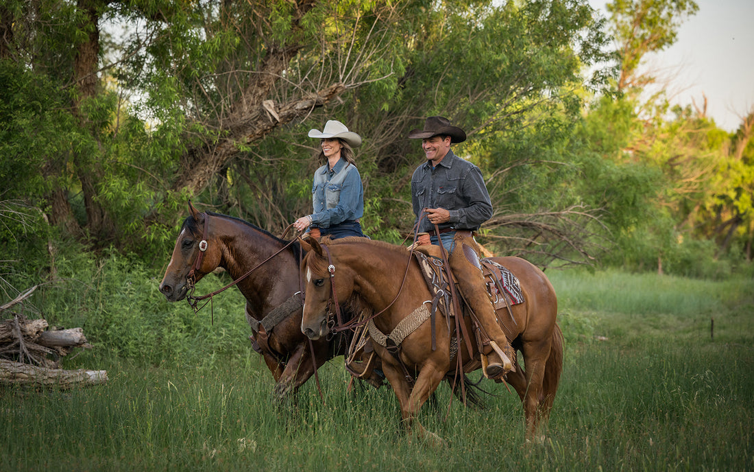 Stran and Jen Smith wearing denim shirts and cowboy hats riding brown horses through a grassy field with trees in the background.