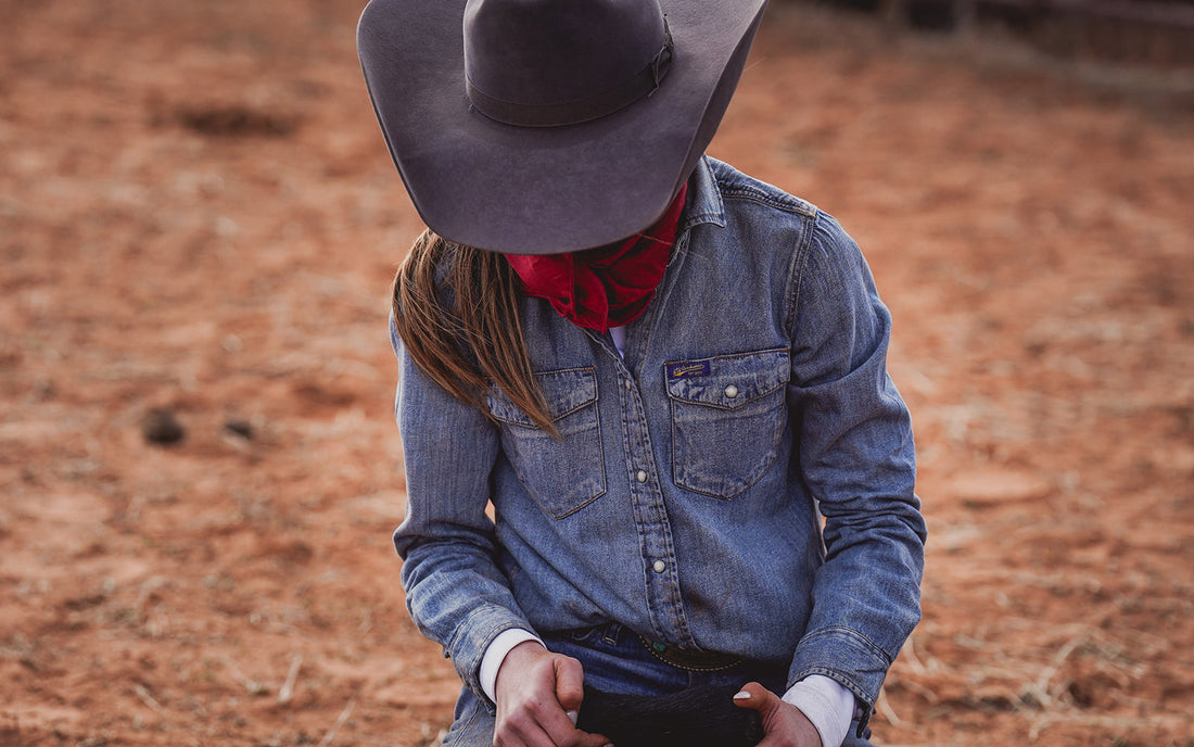 Person wearing a black cowboy hat and blue pearl snap denim shirt standing in a dirt field.