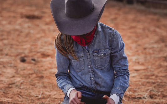 Person wearing a black cowboy hat and blue pearl snap denim shirt standing in a dirt field.
