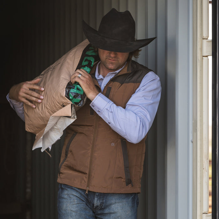 Stetson Smith wearing our two-toned brown vest with double zip closure and classic western yoke accents carrying a bag of feed in front of metal paneling.