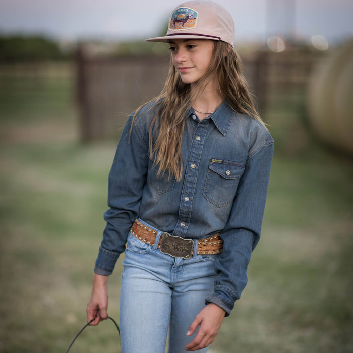 Young lady wearing a tan hat, blue jeans and denim button up shirt walking through a field carrying a bucket.