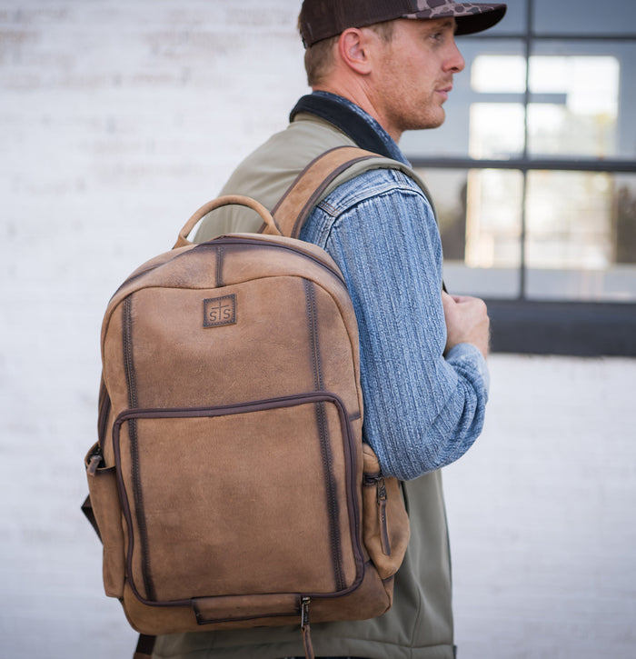 Man wearing an olive vest and long sleeve denim shirt standing in front of a building with a brown genuine leather backpack over his right shoulder.