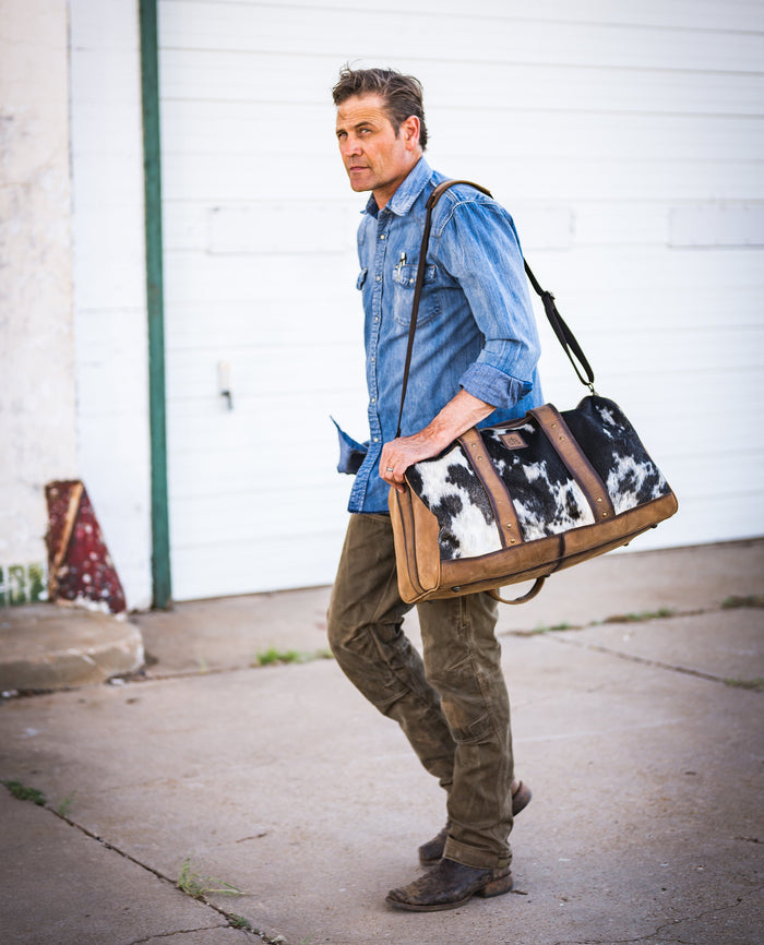 Man wearing khakis and a denim shirt carrying a hair on hide cowhide duffle bag over his shoulder on a sidewalk.