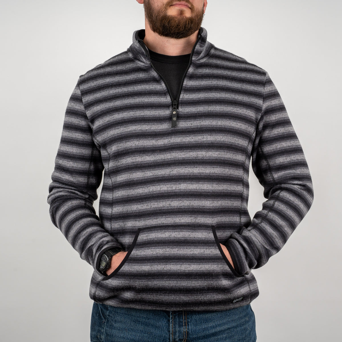 Man standing in front of a gray background wearing a men's quarter zip pullover in black and gray stripes, with his hands in the kangaroo pocket.