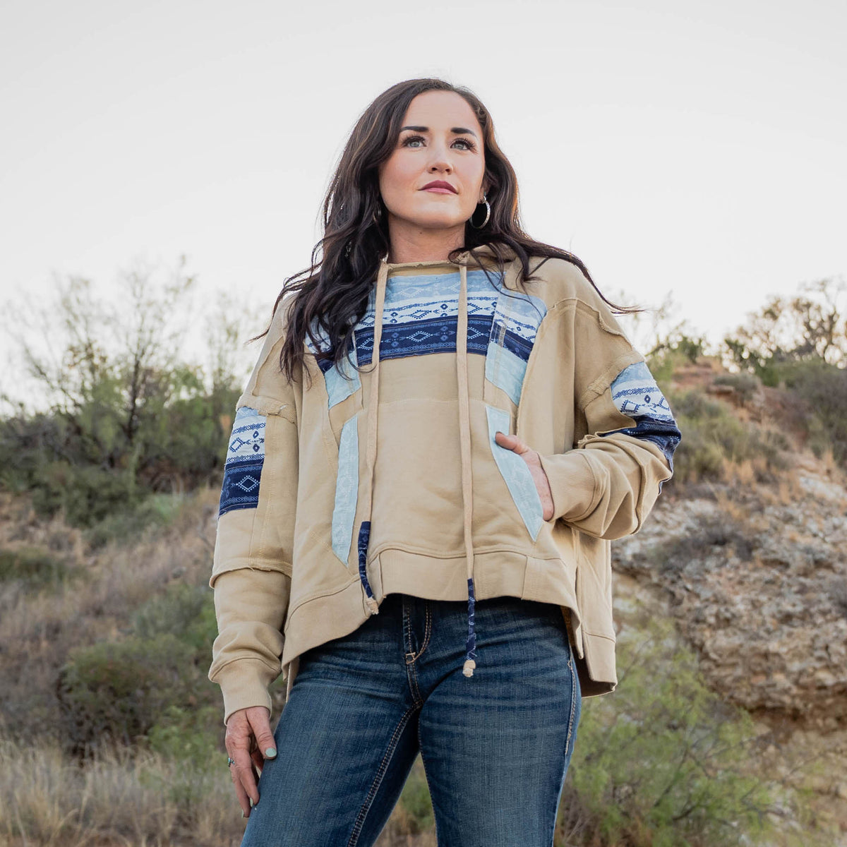 Woman wearing a beige jacket with blue and white patterned sleeves in a desert-like setting.