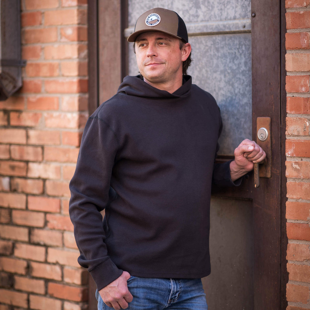 Man leaning against a door wearing a brown hat, jeans and black waffle knit hoodie.