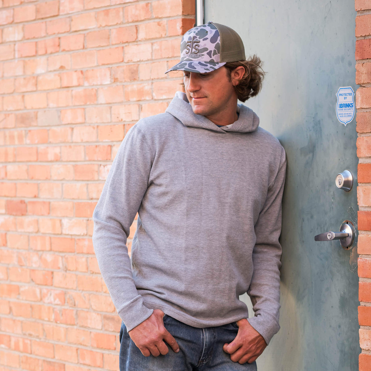 Man leaning against a door wearing a camouflage hat, jeans and gray hoodie. 