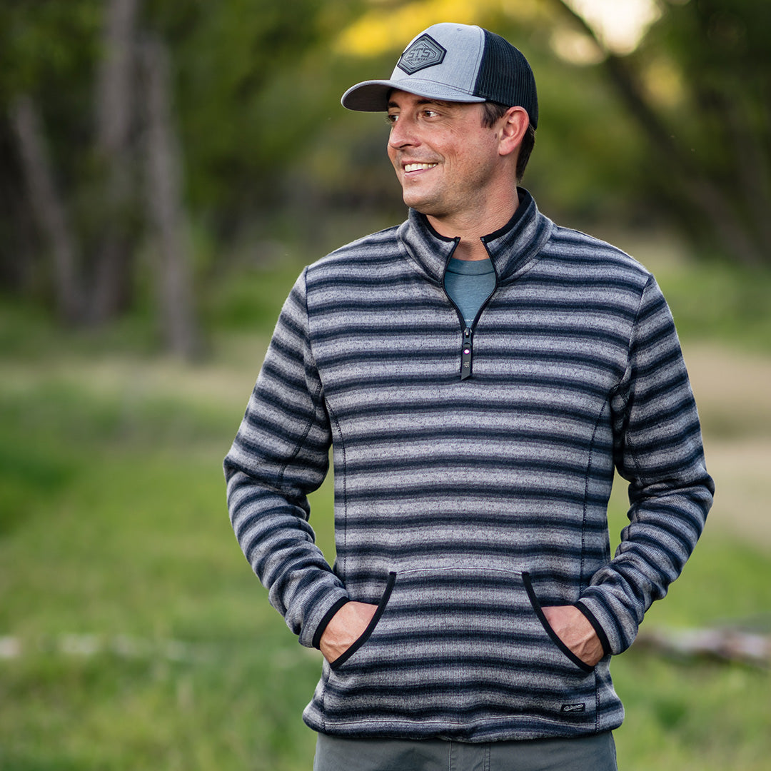 Man standing in the woods wearing a gray hat and men's quarter zip pullover in black and gray stripes, with his hands in the kangaroo pocket.