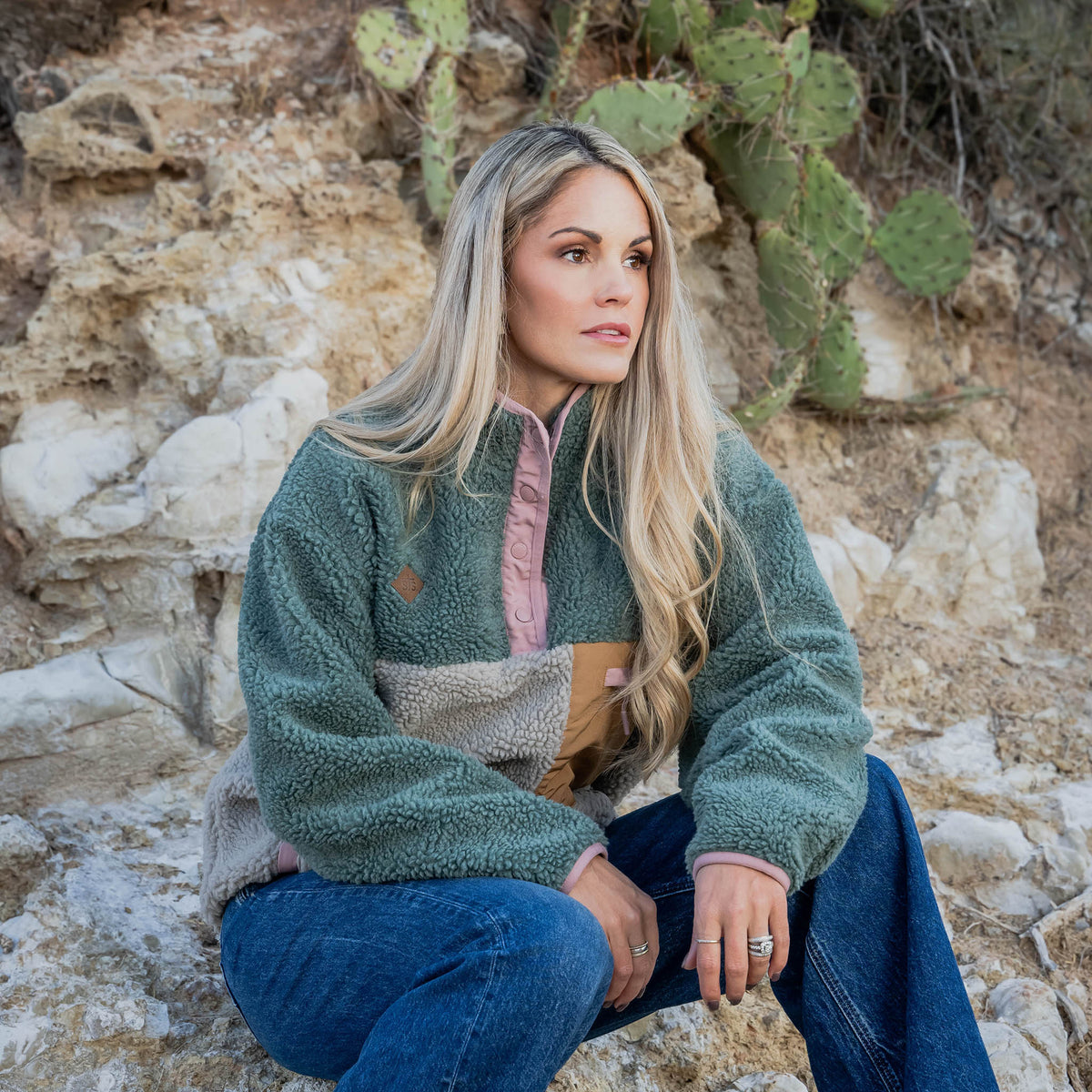 Woman wearing a high plush fleece pullover sitting on rocks with cacti in the background.