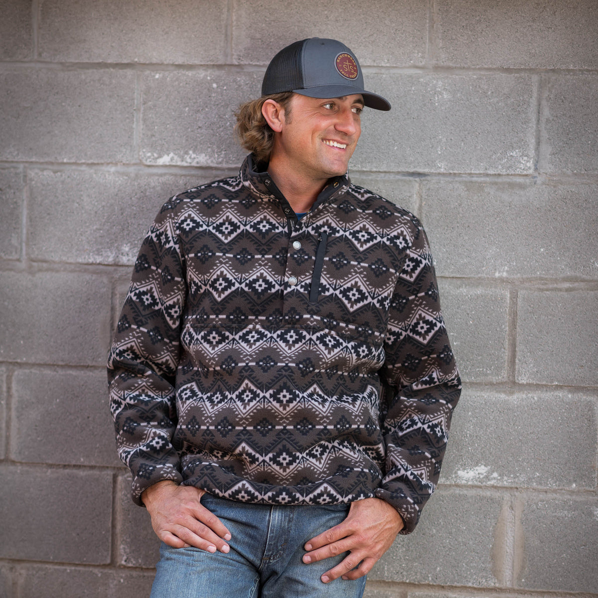 Man leaning against wall wearing gray hat, jeans and men's pullover sweater with a geometric pattern in black, brown, and white colors.