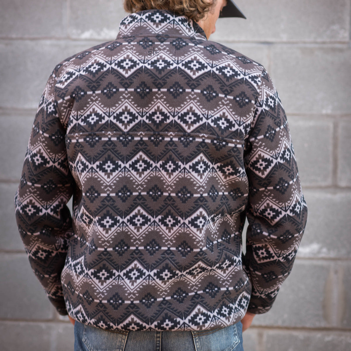 Man facing away in front of wall wearing jeans and men's pullover sweater with a geometric pattern in black, brown, and white colors.
