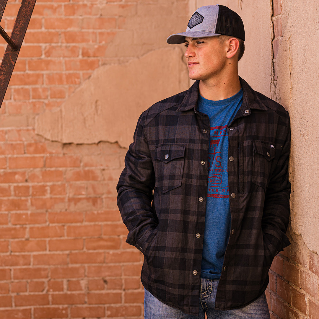 Man wearing a gray hat and long-sleeved black plaid men's shirt jacket with hand warming pockets and snap closures leaning against a brick wall.