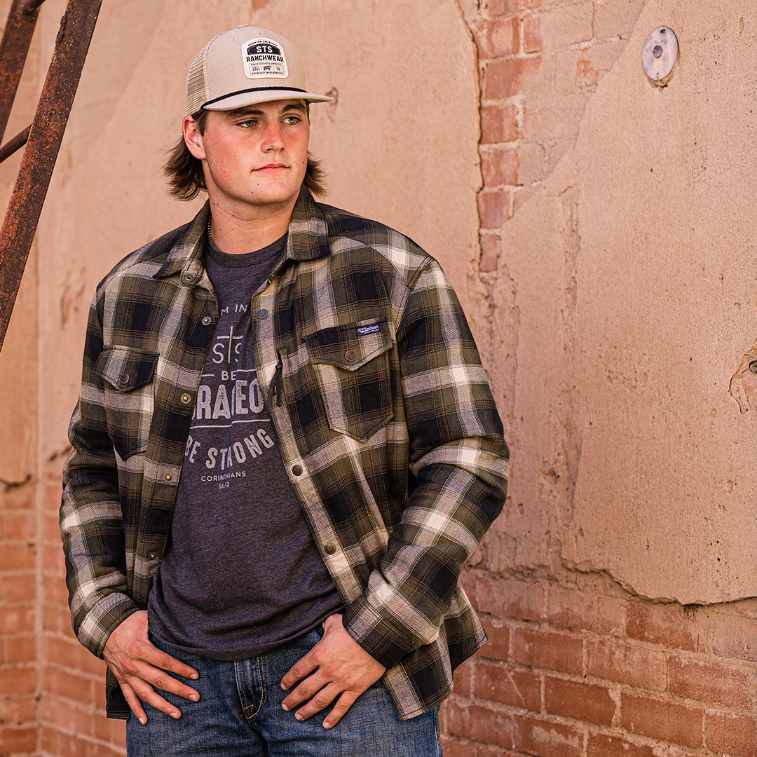 Man wearing a tan hat and long-sleeved plaid men's shirt jacket with hand warming pockets and snap closures in front of a brick wall.