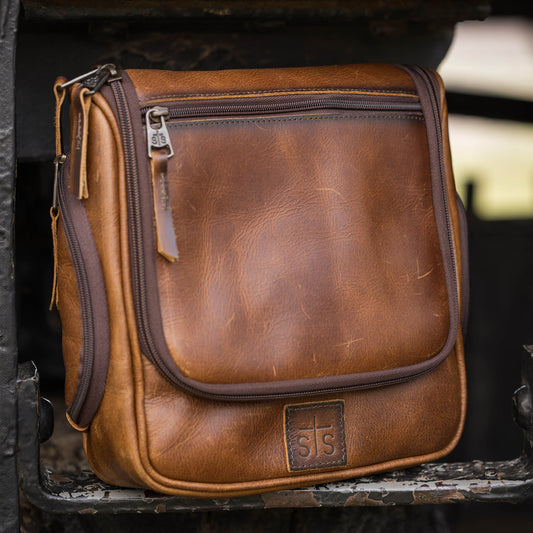 A brown full grain leather toiletry bag with zip closure and a hanging hook, featuring the STS brand logo embossed on a brown leather patch.